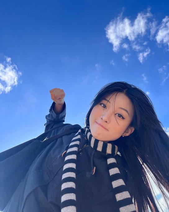 A low-angle selfie captures a woman with long dark hair and a black-and-white striped scarf raising her fist in a cheerful gesture against a bright blue sky, perfectly aligning with the "gm CT happy Sunday" caption.