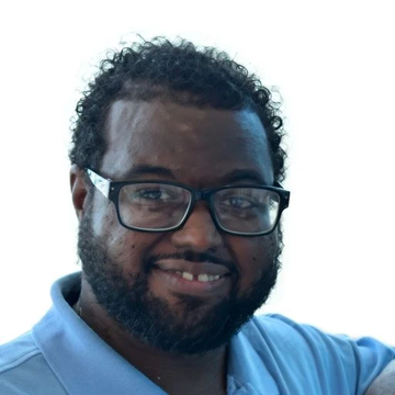 A headshot of a smiling man with curly black hair, a beard, and black-framed glasses, wearing a light blue polo shirt.