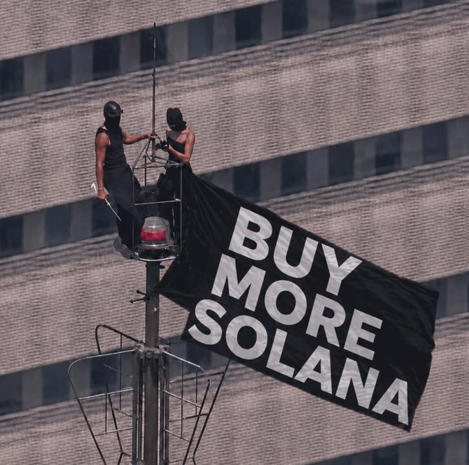 Two masked activists scale a building-mounted pole to unfurl a large black flag bearing the bold white text "BUY MORE SOLANA," echoing the post's "Follow the signs" message as a literal signal urging viewers to purchase the SOL cryptocurrency. The dramatic stunt against a concrete backdrop emphasizes the urgent, attention-grabbing nature of the crypto promotion.
