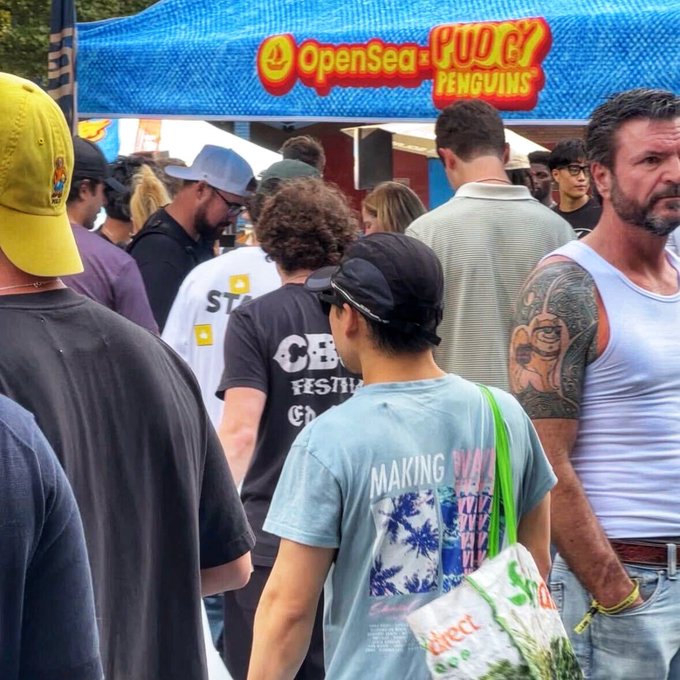 A crowd of NFT enthusiasts gathers at the Pudgy Penguins booth under a blue OpenSea banner, wrapping up Collector Park with branded merch and casual streetwear in tow.