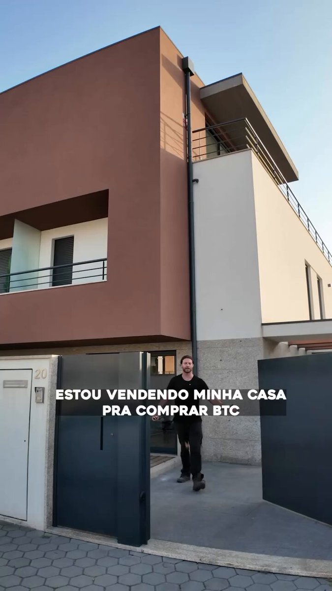 The image shows a man walking through an entrance gate of a large, modern building with a brown facade. The man appears to be dressed in business attire, possibly indicating that he is a real estate agent or involved in property management. The caption on the image reads "Eliezer Tymniak of the infinite via, put one of his houses in Portugal up for sale and said that the intention is to put the entire value into bitcoin:native I think this house is going to turn into about 3 in about 2 years". This suggests that the man might be discussing or promoting a property listing on social media platforms like Facebook.