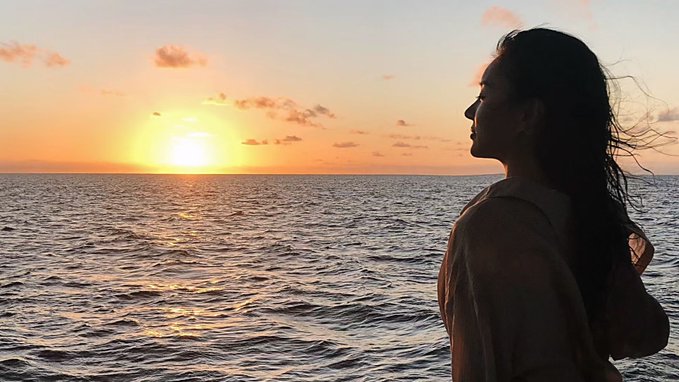 A woman silhouetted against a stunning ocean sunset gazes contemplatively toward the horizon, the warm golden light reflecting on the water's rippling surface.

The image mirrors the introspective tone of the reflection on returning to one's roots in crypto—the peaceful sunset evoking a moment of quiet renewal and reconnection with the original spark of belief in technology's power to change things.