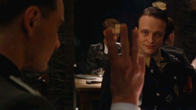The image shows a man wearing a military uniform, with his hand raised in a peace sign gesture towards another person who is also dressed in a military uniform. The scene takes place at an indoor dining table where several people are seated around it. The overall atmosphere of the image suggests that they might be enjoying a meal together or celebrating a special occasion.