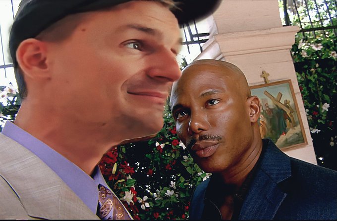 Two men pose for a close-up selfie, one in a suit and cap and the other in a dark shirt, with floral arrangements and a religious crucifixion painting visible behind them.
