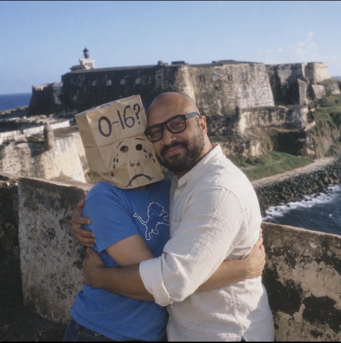 Two men embrace in front of a historic coastal fortress; one man wears a blue shirt and has a paper bag over his head with '0-16?' and a sad face drawn on it, while the other wears glasses and a white shirt.