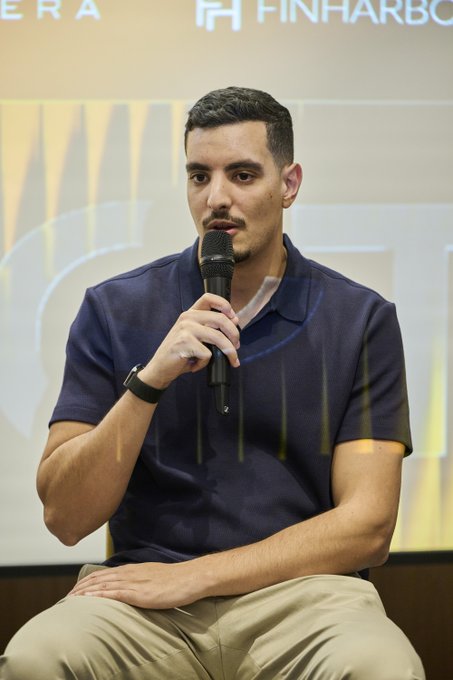 A young man with dark hair and a goatee, wearing a navy blue polo shirt and khaki pants, speaks into a microphone while seated on stage. Behind him, partial logos and the word "FINHABO" are visible, suggesting a professional event setting related to the financial panel discussion mentioned in the caption.