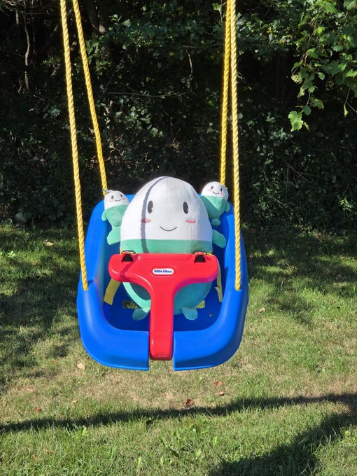 A blue plastic children's swing with yellow ropes, featuring a Little Tikes caterpillar/turtle character design, hanging over grass with trees in the background.