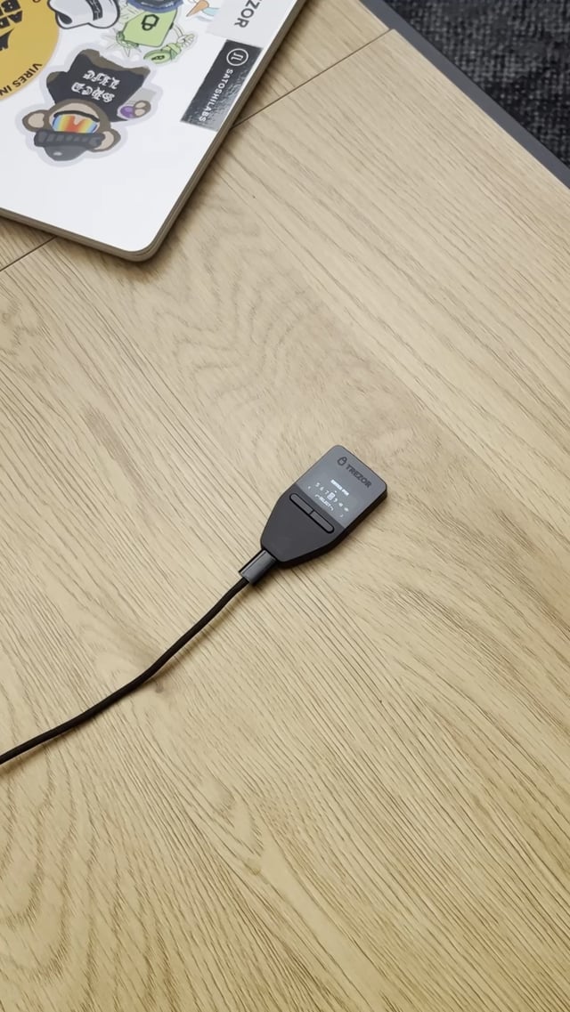 A Trezor hardware wallet rests on a light wooden surface next to a sticker-covered laptop, with a cable attached to it. The image illustrates the article's question about choosing an affordable hardware wallet under $100, as Trezor is one of the popular options in that price range.