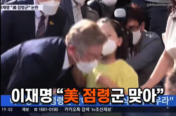 A Korean news broadcast screenshot from @bitcoinvaccine showing a man in a mask, with the headline '이재명 '美 점령군 맞아'' about Lee Jae-myung.