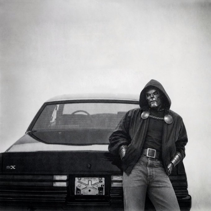 A man wearing a skull mask and hooded jacket leans against the back of a black car with a "GT-X" badge, while a novelty license plate reading "ANOTHER DAY ANOTHER DOUGH" is visible below. The eerie, masked figure creates a dramatic juxtaposition with the casual "Gm Touch Grass today" post, suggesting a tongue-in-cheek, dark-humored reminder to step away from the screen and enjoy the outdoors.
