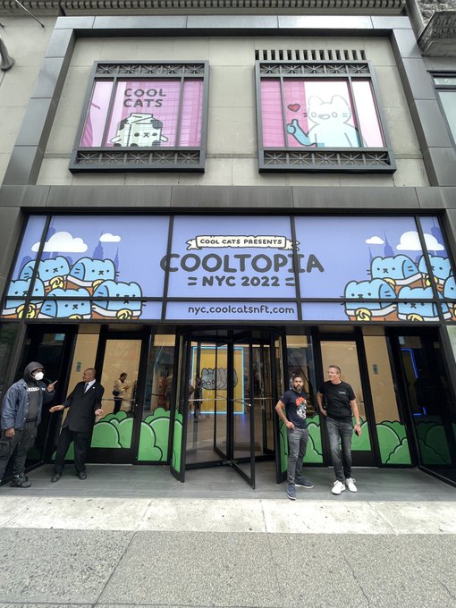 The entrance to a 'COOLTOPIA NYC 2022' pop-up event hosted by Cool Cats at nyc.coolcatsnft.com, featuring cat-themed window displays and two men standing by the revolving glass doors.