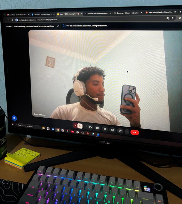 A computer monitor displays a Google Meet session where a man with curly hair and headphones is taking a mirror selfie with his phone, with a colorful backlit keyboard visible on the desk below.