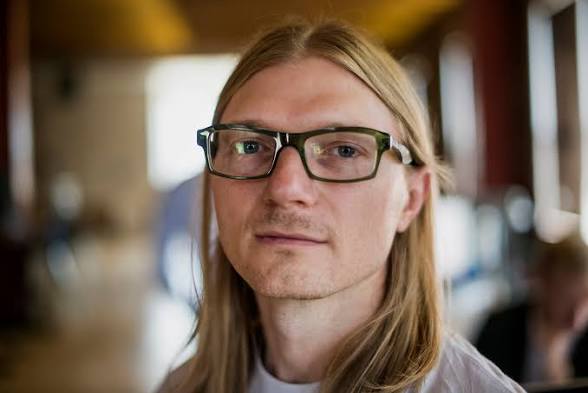 The image shows a young man with long blonde hair and dark-rimmed glasses looking directly at the camera in a softly blurred indoor setting. This appears to be Jesse Powell, the philosophy graduate turned crypto entrepreneur, as referenced in the post about his pivotal 2011 visit to Mt. Gox in Tokyo that launched his journey from selling in-game currency to founding Kraken, which recently sold to Nasdaq at a $21 billion valuation.