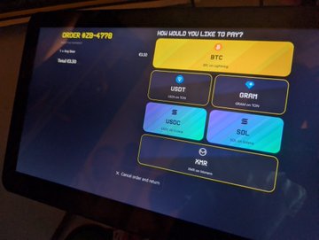 A touchscreen ordering kiosk displaying 'ORDER #29-1778' with a menu of cryptocurrency payment options including BTC, USDT, DAI, USDC, SOL, and XQR.