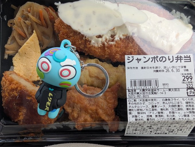 A Japanese bento (ジャンボのり弁当) containing fried foods, rice, and sides, with a blue character keychain attached and a label showing 299 (税込322) yen.