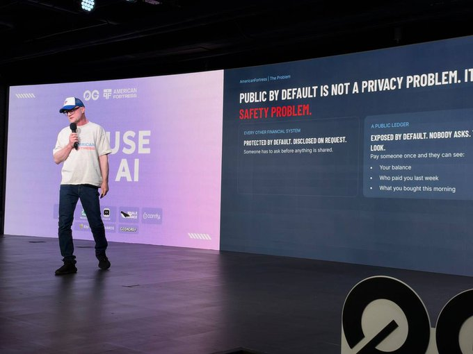 A man in a white t-shirt and cap speaks on stage, with a presentation slide beside him reading 'PUBLIC BY DEFAULT IS NOT A PRIVACY PROBLEM. IT'S A SAFETY PROBLEM' and explaining that financial information is protected by default unless disclosed on request.