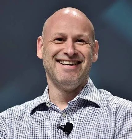 A bald, smiling man in a checkered button-down shirt speaks at what appears to be a conference or interview.