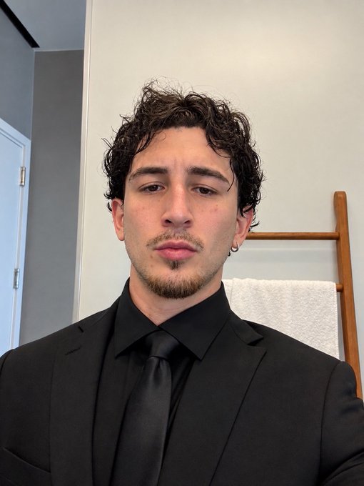 A young man with curly dark hair, a small mustache, and a goatee wears a black shirt, black tie, and black suit jacket indoors.