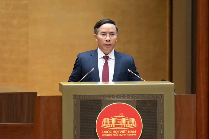 A man in a dark suit and red tie speaks at a podium labeled 'QUỐC HỘI VIỆT NAM / NATIONAL ASSEMBLY OF VIET NAM'.