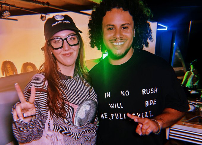 Two people pose together smiling for the camera; a woman with long red hair, glasses, and a black cap flashes a peace sign, while a man with curly hair wears a black t-shirt reading 'IN NO RUSH WILL RIDE [CAREFULLY]', illuminated by green stage lighting.