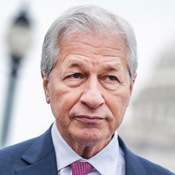 A distinguished older man with gray hair, wearing a navy suit, white shirt, and burgundy patterned tie, looks directly at the camera with a serious expression against a softly blurred background. The image accompanies news of JPMorgan CEO Jamie Dimon's warning about the US potentially losing its reserve currency status within 25 years, suggesting the man pictured is Dimon himself.