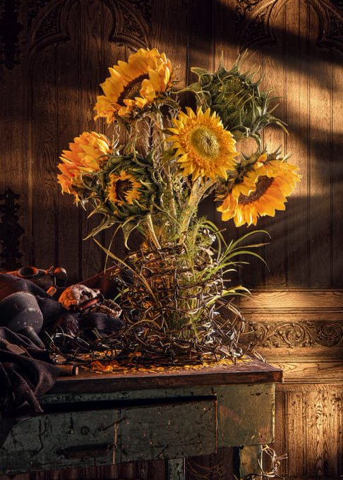 A moody still life photograph of a bouquet of sunflowers in a glass vase on a distressed wooden table, illuminated by warm shafts of light against a dark, carved wooden background.