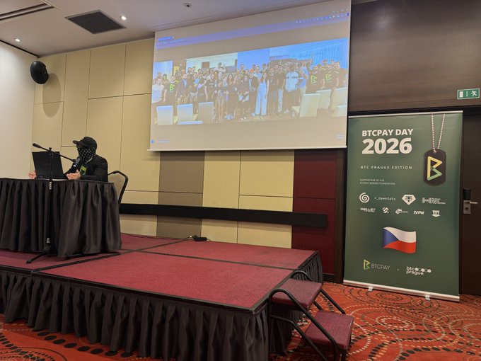 The image shows a stage with a man standing behind a podium, giving a presentation or speech at an event called "BTCPay Day". The stage has a red carpet covering its floor, adding a touch of elegance to the setting. A large screen is visible in front of the speaker, displaying information related to the event. There are several people present on the stage and around it, indicating that this is a public gathering or presentation.