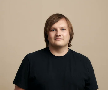 A portrait photo of a man with medium-length brown hair wearing a black t-shirt against a beige background.