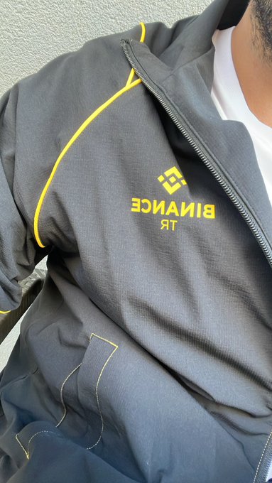 A close-up of a gray 'BINANCE TR' branded jacket with yellow trim, worn over a white shirt.
