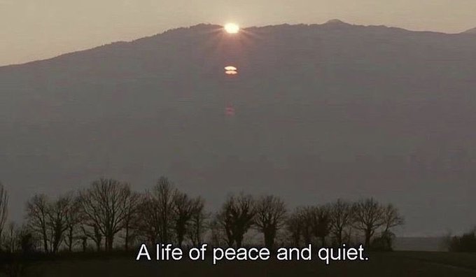 A misty landscape at sunset with the sun setting behind a mountain ridge above silhouetted trees, captioned 'A life of peace and quiet.'
