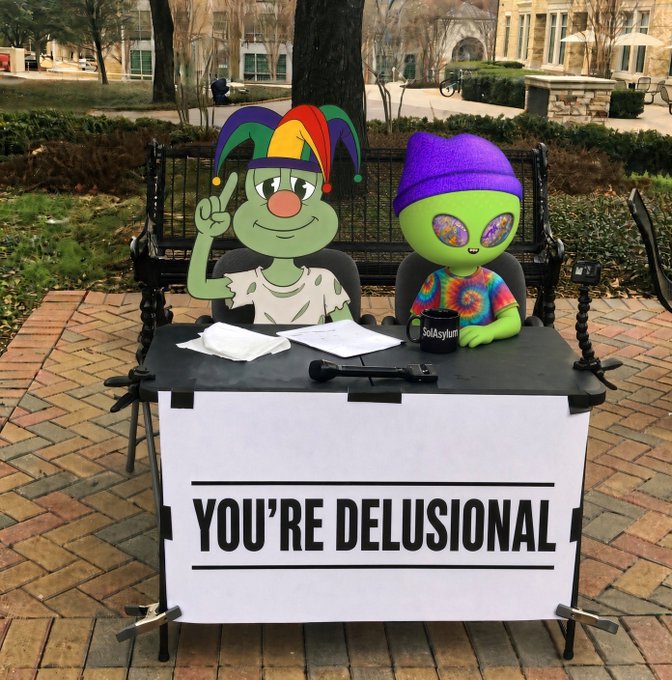 Two cartoonish figures—one a green jester-like character and the other a green alien in a purple beanie and tie-dye shirt—sit behind a table with a sign reading 'YOU'RE DELUSIONAL' at an outdoor campus event.
