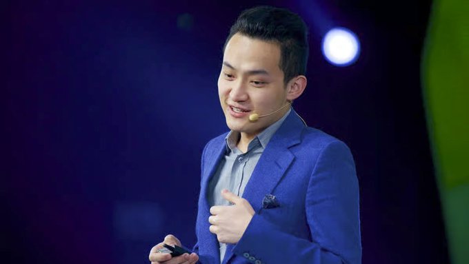 The image features a man wearing a blue suit standing on stage with his hands at his sides, giving him an air of confidence as he speaks into a microphone. The background shows a dark purple color, which contrasts with the white wall behind him. There is also a green plant visible in the scene. The post that accompanies this image reads: "Justin Sun's Long-Term Strategy: Building a Global Blockchain Ecosystem In the rapidly changing crypto industry, long-term strategies are often more important than short-term trends." This statement seems to be related to the man speaking at the event and his vision for the future of blockchain technology.