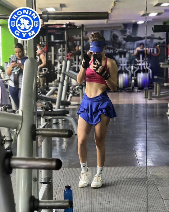A woman in a pink sports bra, blue skirt, blue visor, and fingerless gloves takes a mirror selfie in a gym, with a 'RONIN GYM' logo in the top left corner.