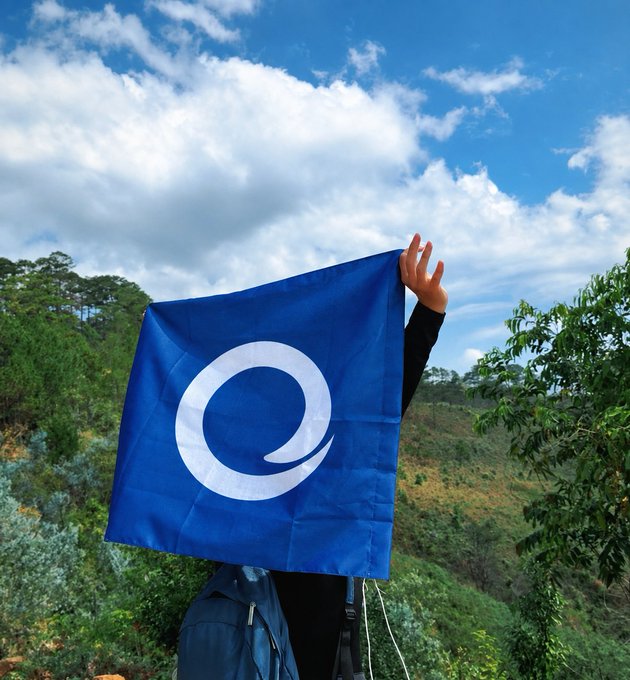 A hiker holds up a blue flag featuring a white spiral logo against a scenic mountainous backdrop, embodying the post's message: by venturing outdoors into the wilderness, they help keep internet services running for those who stay connected online.