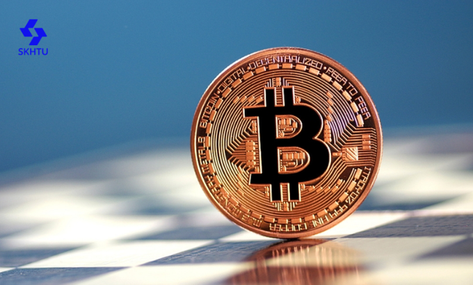 A copper-colored physical Bitcoin token featuring the iconic "₿" symbol stands upright on a checkered surface against a blue background, with the "SKHTU" logo visible in the upper left corner. The image visually reinforces the accompanying post about SKHTU Exchange's analysis of Bitcoin's cyclical movements and market structure following a recent pullback.