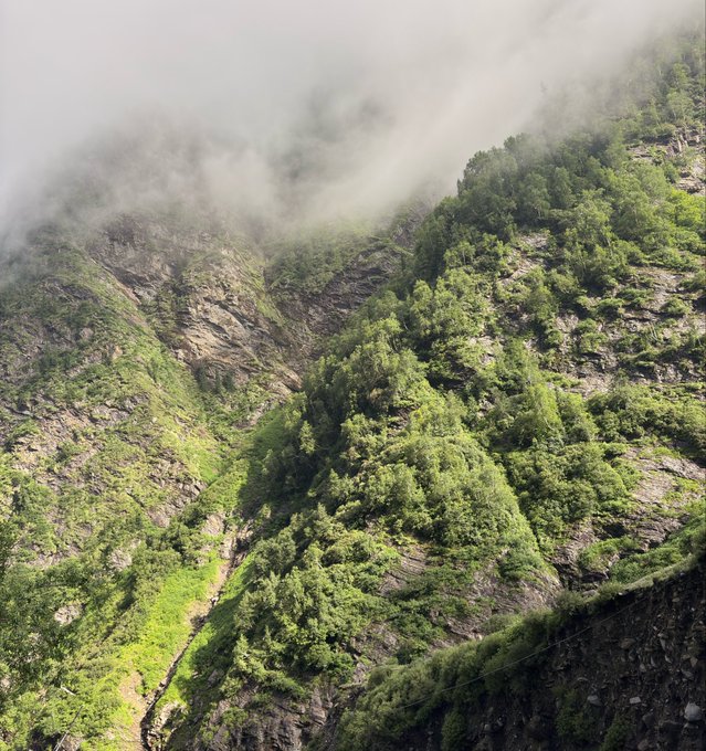 A scenic view of a steep, rocky mountain valley covered in lush green vegetation with misty clouds rolling over the peaks.