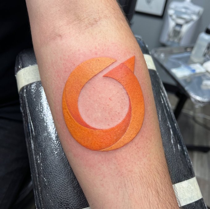 A freshly done orange circular logo tattoo on a person's forearm, sitting on a black armrest.