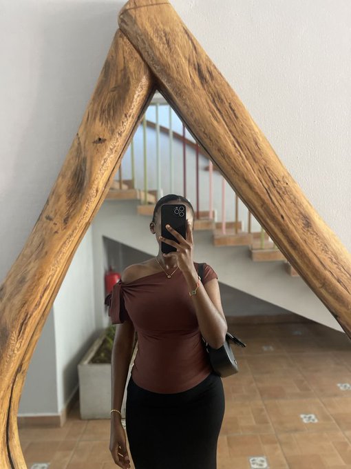 The image features a woman taking a mirror selfie while wearing a brown top, black skirt, and holding a purse. The backdrop includes a wooden wall with a metal staircase visible behind her. The caption of the image reads "good morning beautiful people check out the quoted tweet to see cool shit I'am still focused on $RIVER and I'm dropping a post with a graphic I'm currently on my way to work do you have your day planned out?".