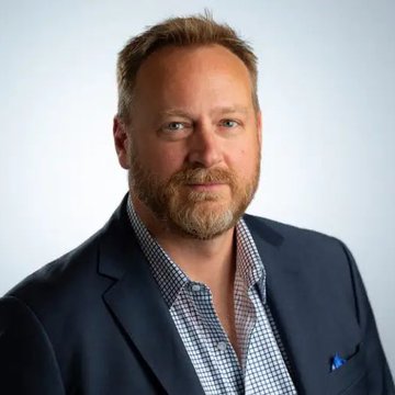 A professional headshot of a man with a beard, wearing a dark blue suit jacket over a checkered shirt with an open collar.
