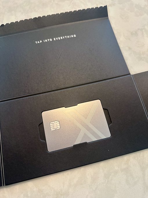 A sleek, silver metal credit or debit card with a geometric design and chip, resting in its black presentation folder which reads 'TAP INTO EVERYTHING.'
