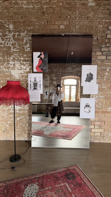 A mirror leans against a weathered brick wall, reflecting a person in a hat taking a photo, surrounded by fashion sketches and a red fringed lamp on a rug.