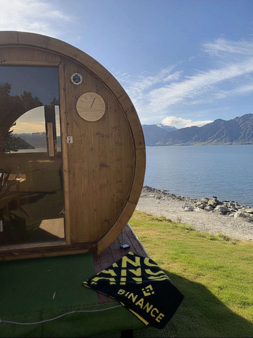 A wooden barrel sauna sits lakeside against a mountainous backdrop, with a "BINANCE" towel visible on the green turf outside, humorously illustrating the post's "offline" theme as the crypto exchange temporarily steps away from its digital trading platform for a literal disconnected retreat.