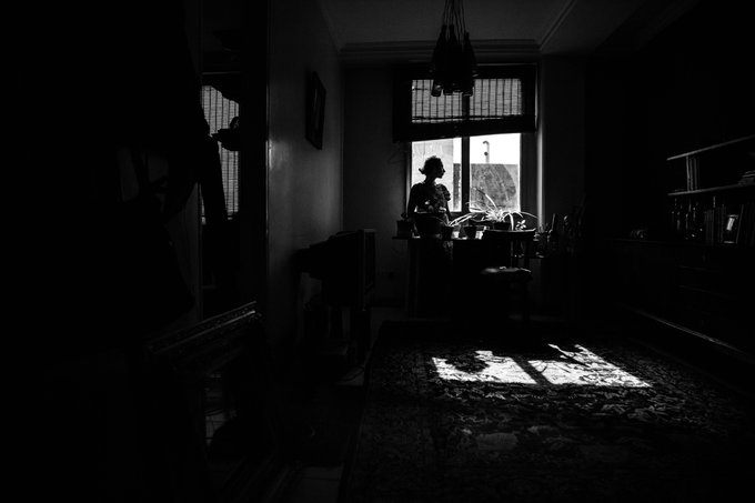 A black and white atmospheric photograph showing a silhouetted figure standing by a window in a dimly lit interior, with light streaming through onto an ornate rug.