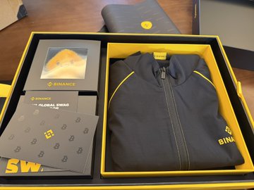A dark Binance-branded zip-up jacket with yellow accents is displayed in an open promotional gift box, alongside cards labeled "BINANCE" and "F1 GLOBAL SWAP." The image showcases the premium quality Binance merchandise referenced in the post, humorously suggesting it as consolation gear for trading losses.