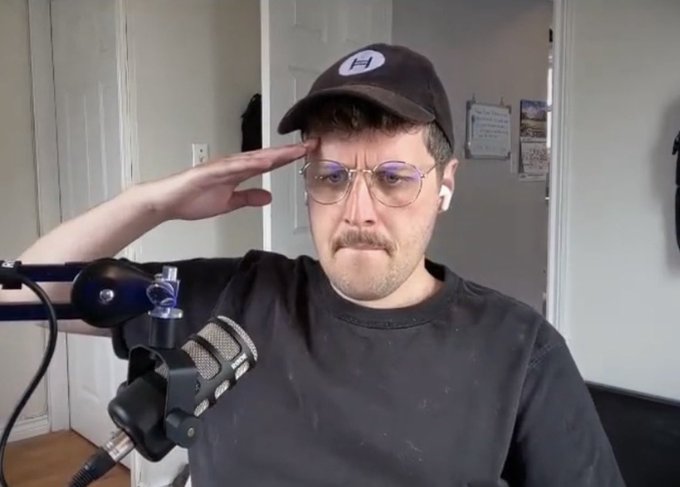 A man wearing a black cap and glasses salutes with a serious expression while sitting in front of a microphone during what appears to be a podcast recording.