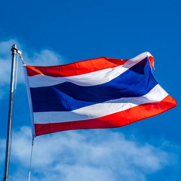 The flag of Thailand billows against a bright blue sky, its distinctive red, white, and blue horizontal stripes clearly visible. The image visually ties to the post about Thailand's SEC implementing new Travel Rule requirements for crypto exchanges, symbolizing the country's regulatory authority over cryptocurrency transactions.