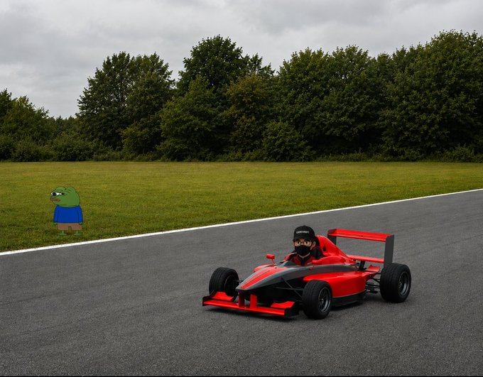 The image features a red race car driving down an asphalt road with trees in the background. A person wearing a blue shirt can be seen standing next to the car, possibly observing or waiting for their turn to drive. The scene suggests that this could be a racetrack where people are participating in racing events or simply enjoying leisurely drives around the area.