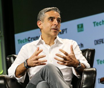 A man in a white shirt and jeans sits in a black leather chair speaking on stage at a TechCrunch event, gesturing with both hands.
