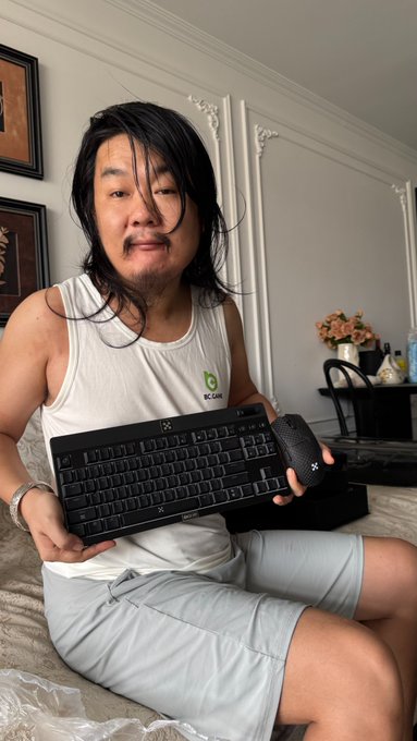 A man with long dark hair, wearing a white tank top and gray shorts, sits on a bed holding a black OKX-branded keyboard and matching mouse. The image accompanies his post thanking OKX for sending the keyboard, which he plans to use for writing tweets.