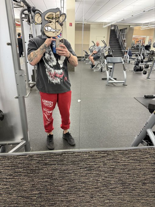 A person wearing a graphic skull t-shirt, red sweatpants, and gray sneakers takes a mirror selfie in a gym, with their face covered by a cartoon monkey sticker.